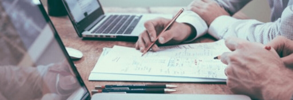 Pen pointing at specific figures of a financial forecast on paper, next to open laptops on a table